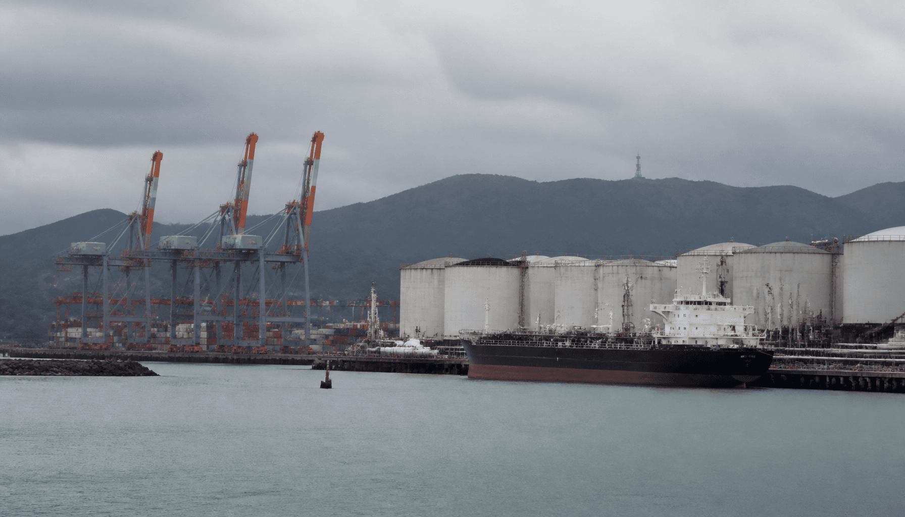 Container cranes and a tanker berthed at a New Zealand port terminal under overcast sky