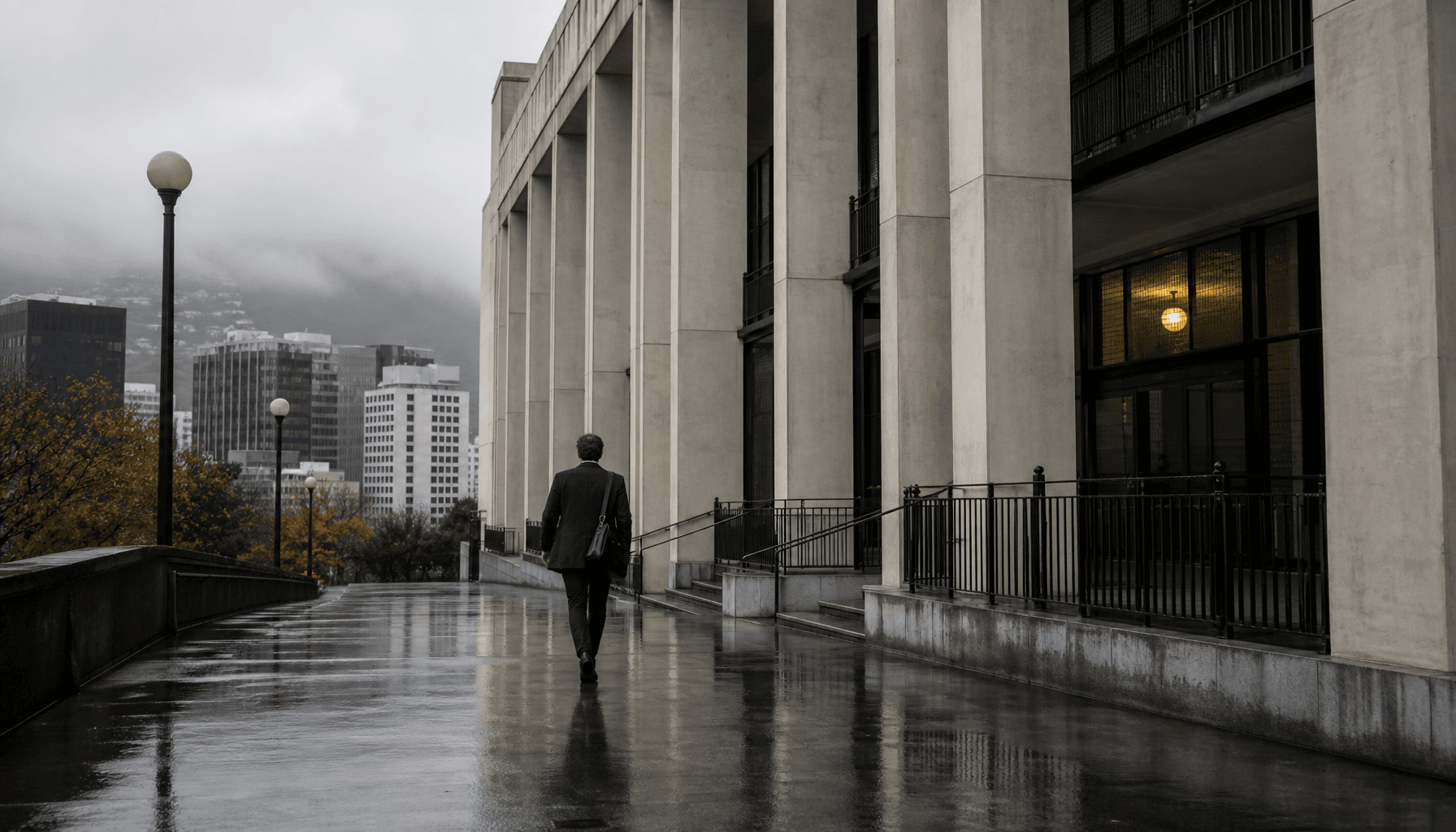 A lone professional walks toward a Wellington civic building on a grey autumn morning, laptop bag in hand