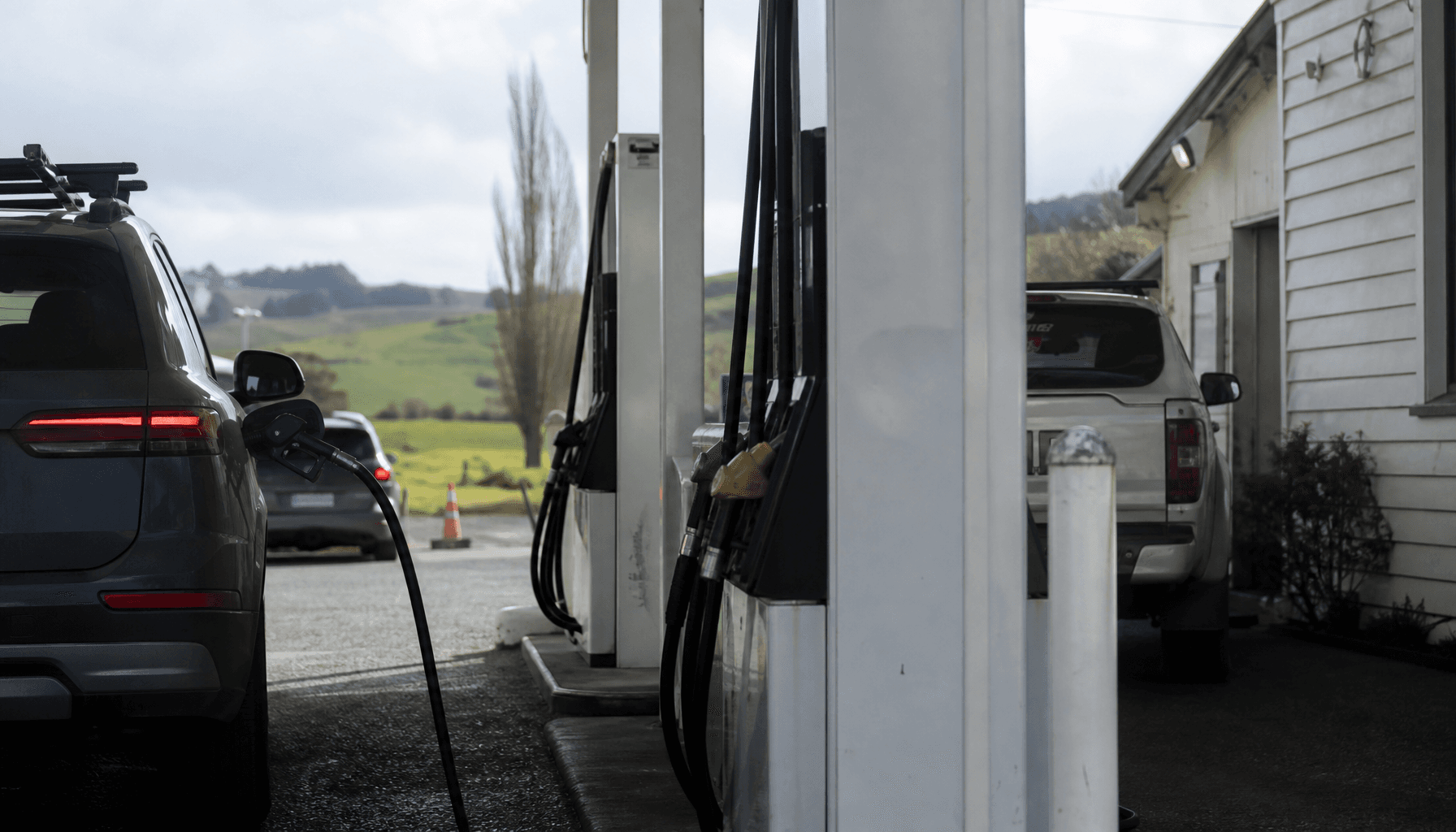 New Zealand regional petrol station forecourt with fuel pumps, overcast midday light, green paddock visible in distance.